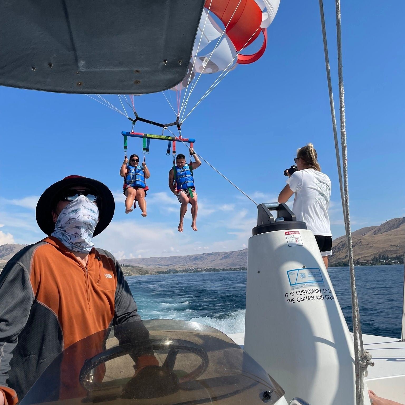 Two people are parasailing over a lake, wearing life jackets, while a boat pulls them. A person drives the boat and another takes photos. Mountains and blue sky are visible in the background.