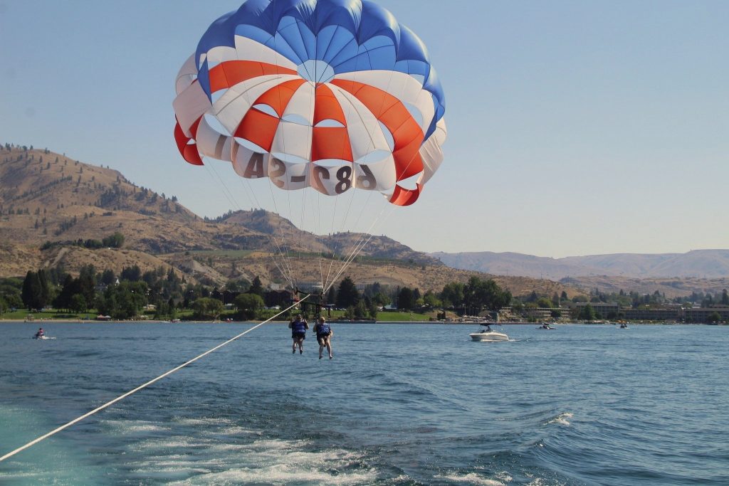 Two people are parasailing over a lake, suspended by a large red, white, and blue parachute. The parachute is attached to a boat speeding across the water, with mountains and trees visible in the background.