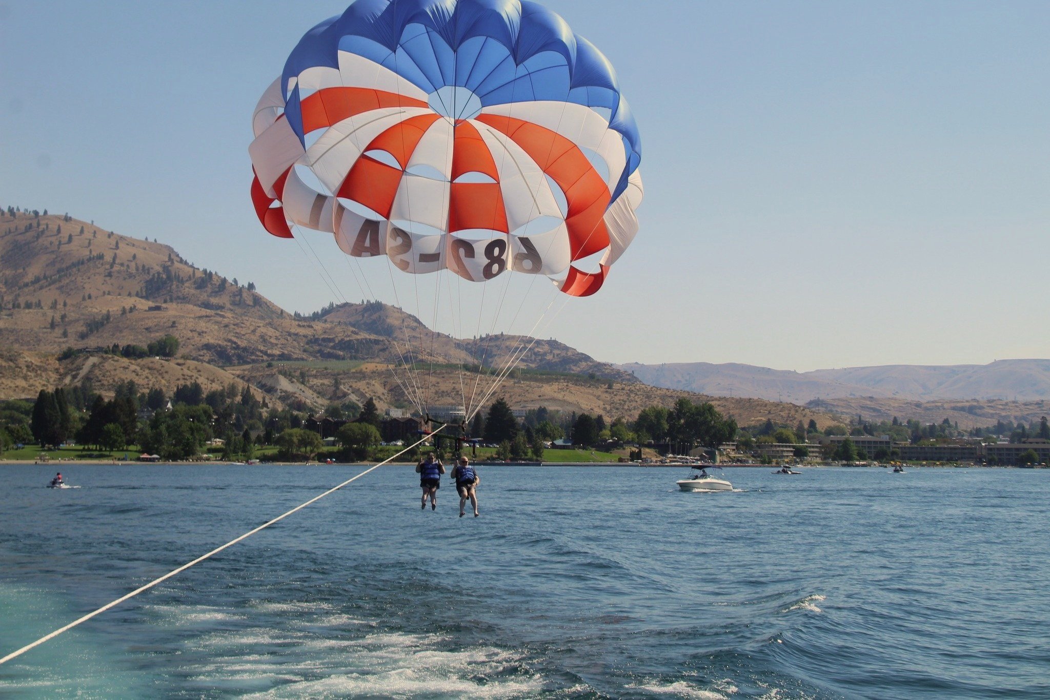Two people are parasailing over a lake, suspended by a large red, white, and blue parachute. The parachute is attached to a boat speeding across the water, with mountains and trees visible in the background.
