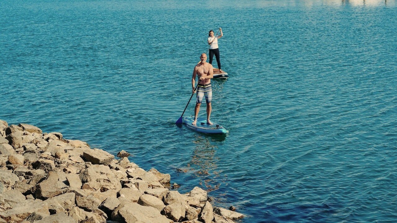 Two people are enjoying a paddle board adventure on calm blue water near a rocky shoreline. One is shirtless and closer to the camera, while the other stands farther back, steadily paddling along the serene scene.